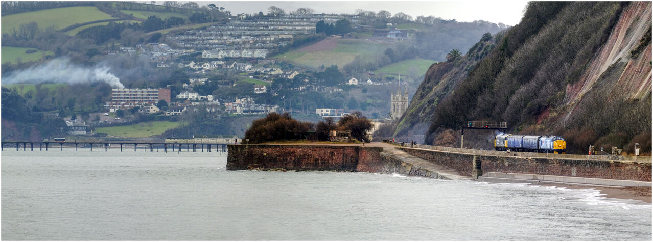 Teeside Traveller
37501 leads 37512 along the sea wall at Teignmouth passing Sprey point. They were returning from Penzance Long Rock depot having delivered a Class 175 unit. 
Despite the grey day i tried a panoramic shot of the area the distant smoke from a bonfire providing me with a little inspiration.
